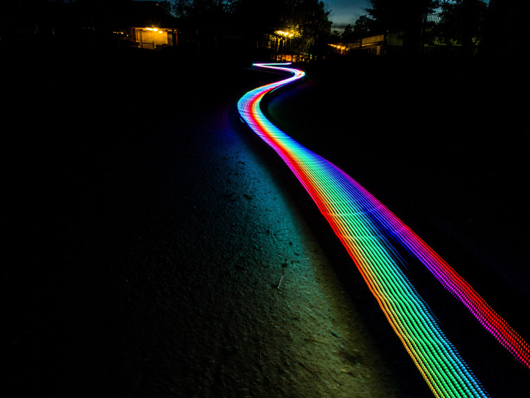 A long-exposure night photo of a winding path with a rainbow-colored light trail.