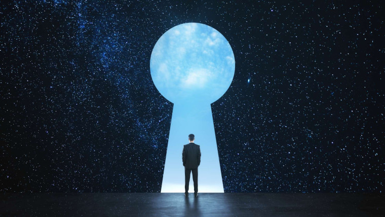 A person in a suit standing before a large keyhole-shaped opening in a starry night sky, looking out toward a bright blue sky.