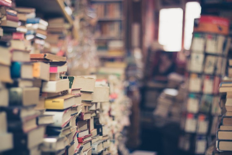 A crowded bookshop with tall stacks of books piled in the foreground and shelves of books receding into the background, with a blurred figure visible in the distance.