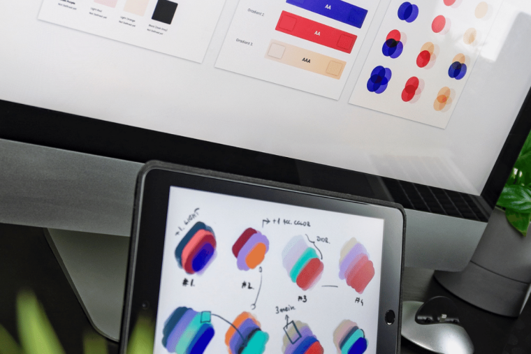 A designer's workspace showing a monitor with a color contrast accessibility guide and a tablet with hand-drawn color palette sketches.
