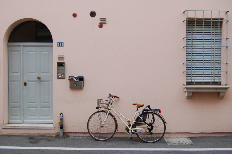 A bicycle parked on a street beside a pink building with an arched doorway and iron-grated window.