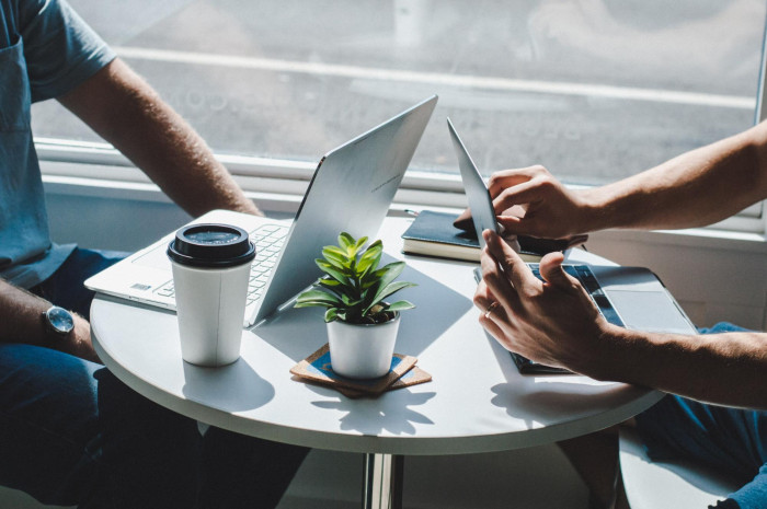 Two people working across from each other at a small round table with laptops, a coffee cup, and a small plant.