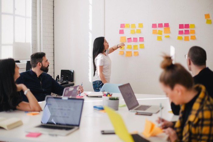 A person leading a team workshop, pointing at colorful sticky notes grouped on a wall while colleagues watch from around a conference table with laptops.