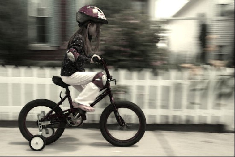 A child wearing a helmet rides a bicycle with training wheels, the background blurred with motion.