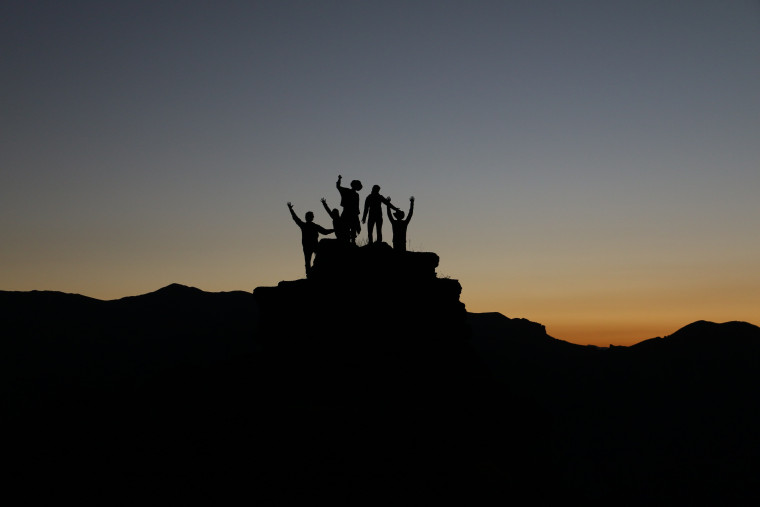 Silhouettes of a group of people with arms raised on a rocky hilltop at sunset.