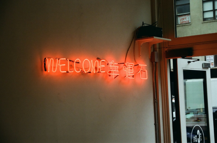 A neon 'Welcome' sign in English and Chinese characters mounted on a wall beside a storefront entrance.