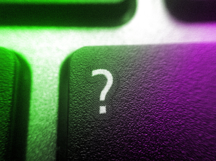 A close-up macro photo of a keyboard's question mark key, lit with green light on one side and purple on the other.