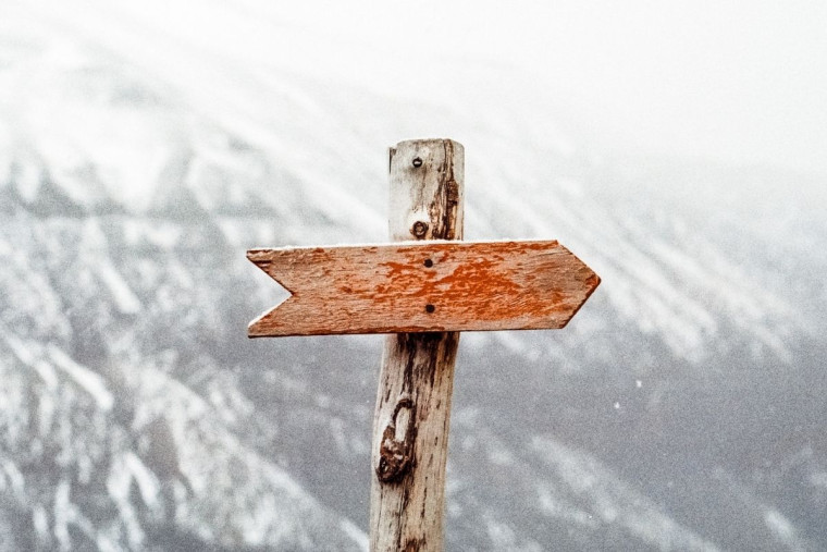 A weathered wooden directional signpost with two blank arrow boards pointing in opposite directions, against a snowy mountain backdrop.