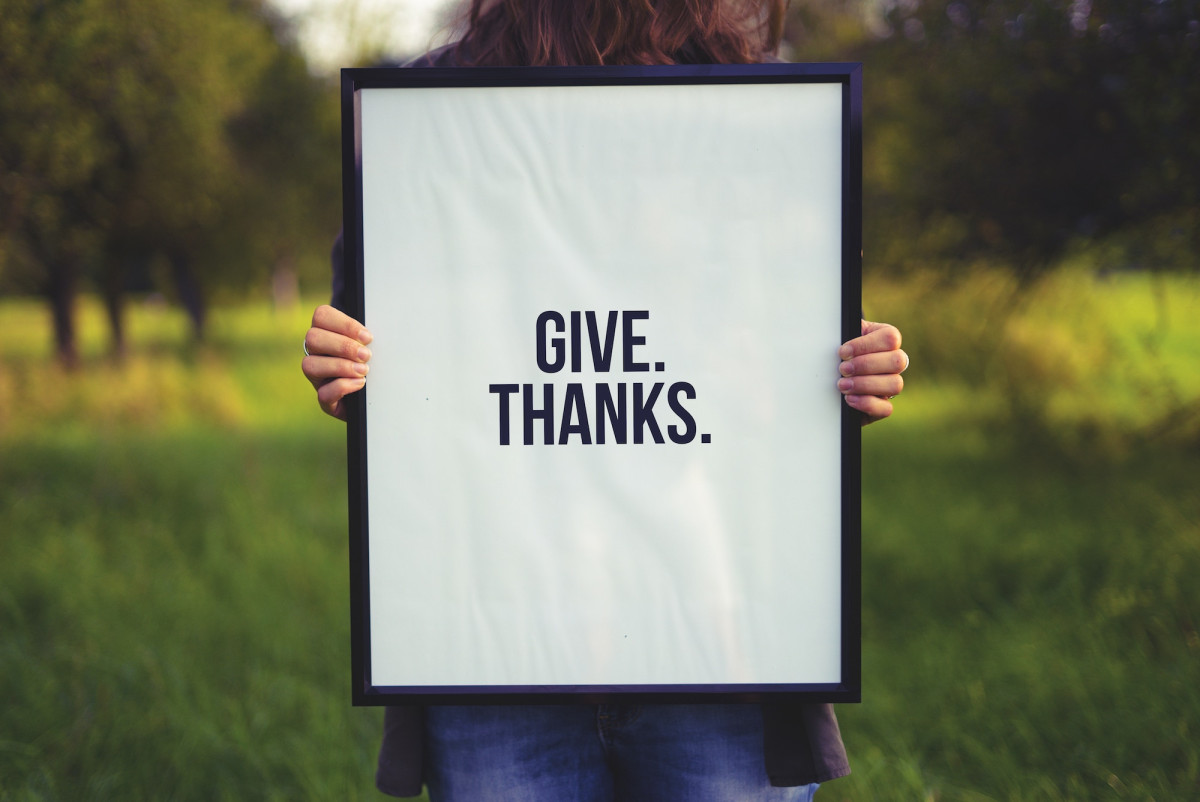 A person holding a framed sign outdoors that reads 'Give. Thanks.'