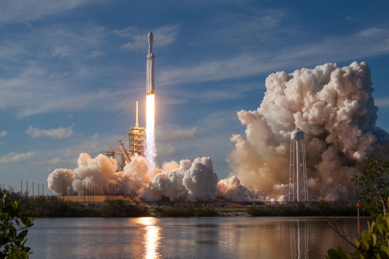 A rocket launching from a seaside pad, trailing a bright flame and billowing clouds of exhaust smoke reflected in the water below.