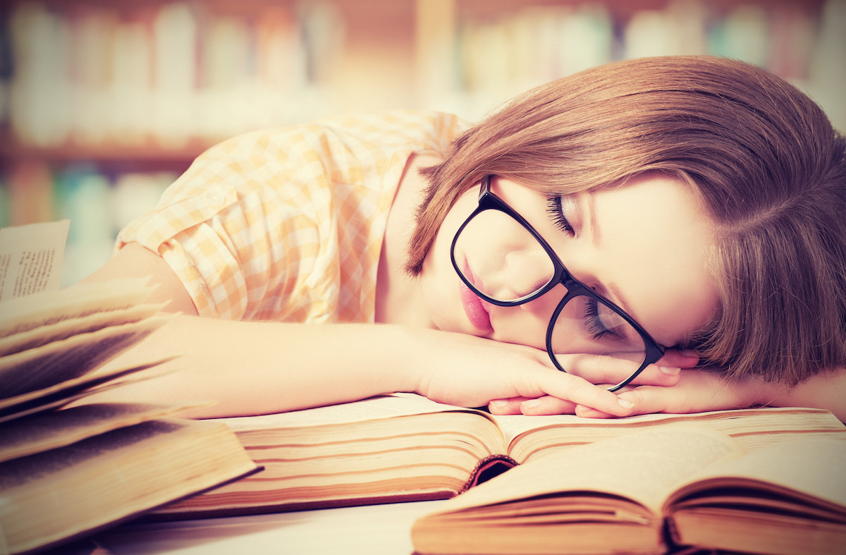 A person wearing glasses asleep on open books in a library
