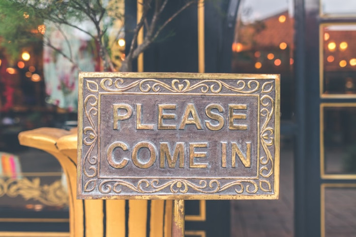 An ornate brass 'Please Come In' sign mounted on a storefront door.