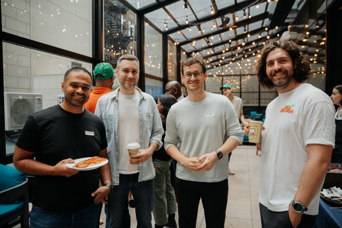 Four attendees posing with coffee and food at a Chameleon networking event in a glass-roofed indoor venue with string lights